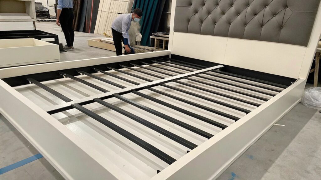 Worker assembling a large bed frame with metal slats and a tufted headboard in a factory setting.