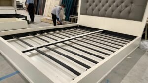 Worker assembling a large bed frame with metal slats and a tufted headboard in a factory setting.