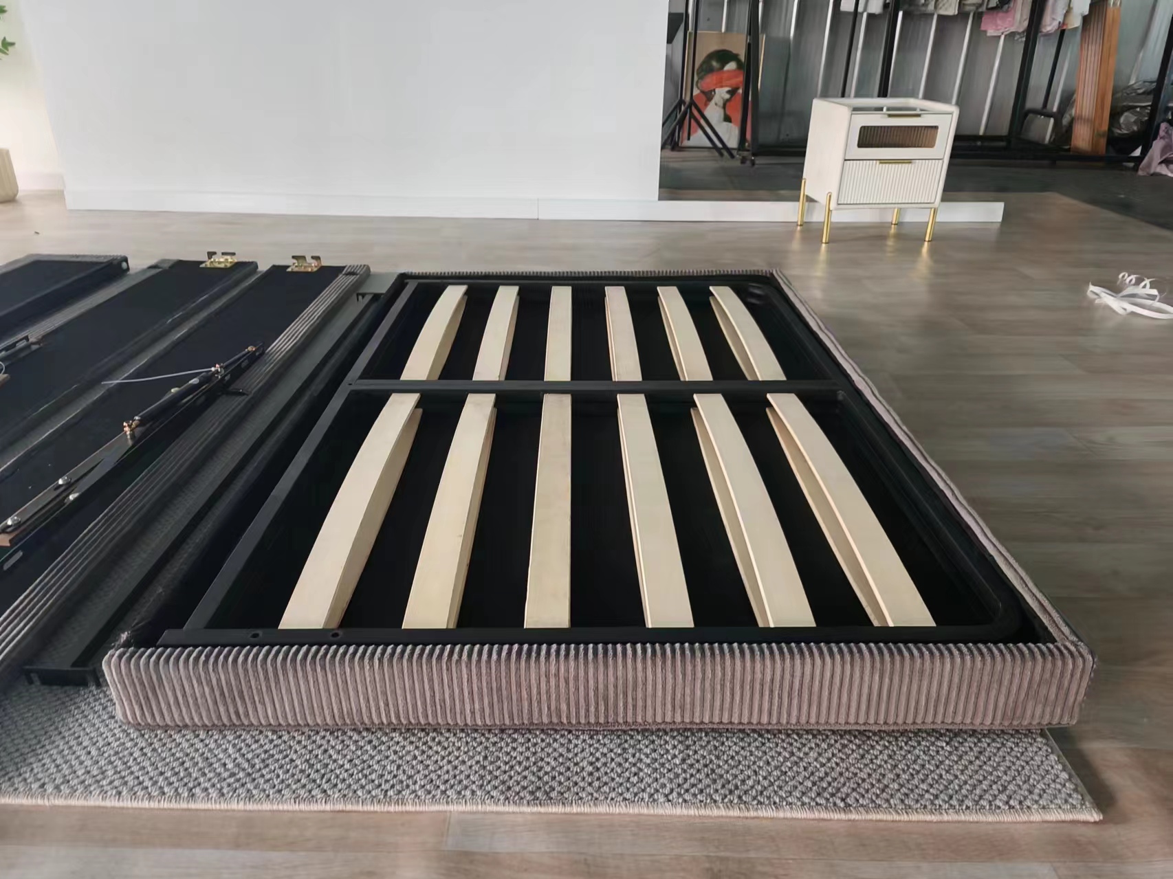 Foldable upholstered bed frame with ribbed fabric design, metal structure, and wooden slats, displayed on a rug in a showroom.