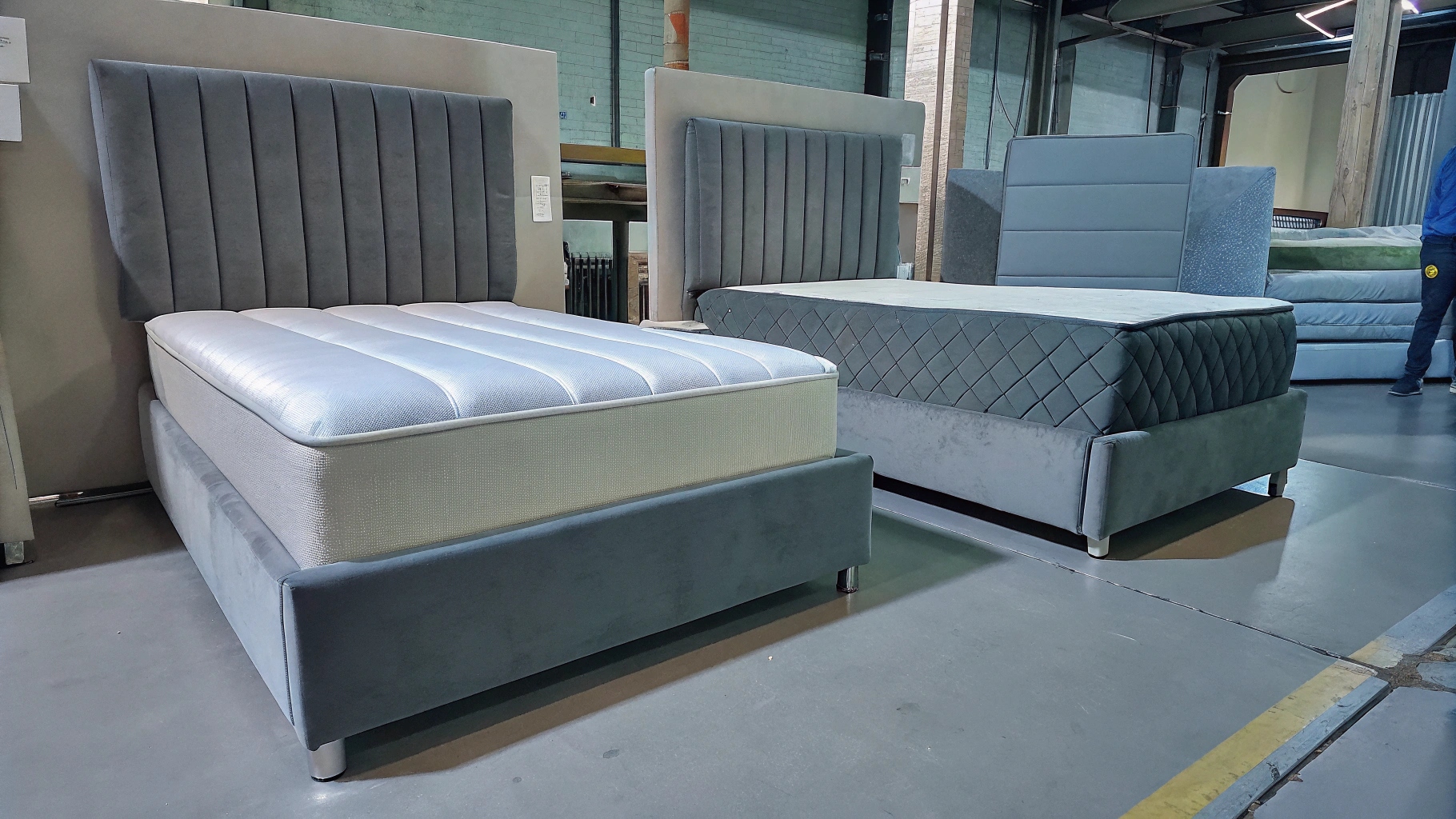 Two modern upholstered beds with grey velvet headboards and mattresses, displayed in a showroom. One bed features a vertical channel tufted design, while the other has a diamond-patterned headboard.