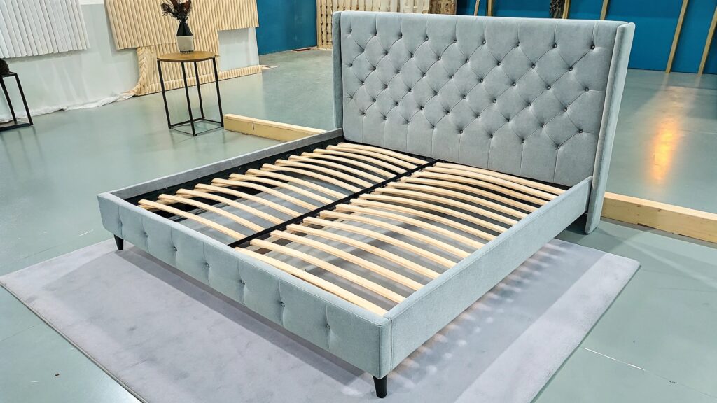 A modern upholstered bed frame in light grey with a tufted headboard, featuring wooden slats for support, displayed in a showroom setting.