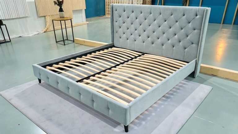 A modern upholstered bed frame in light grey with a tufted headboard, featuring wooden slats for support, displayed in a showroom setting.